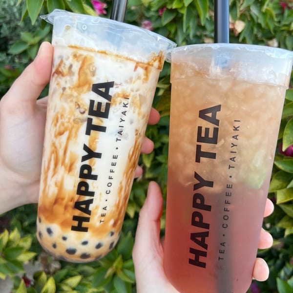 Two hands holding Happy Tea drinks, one brown sugar boba and one iced tea, against a leafy green background.