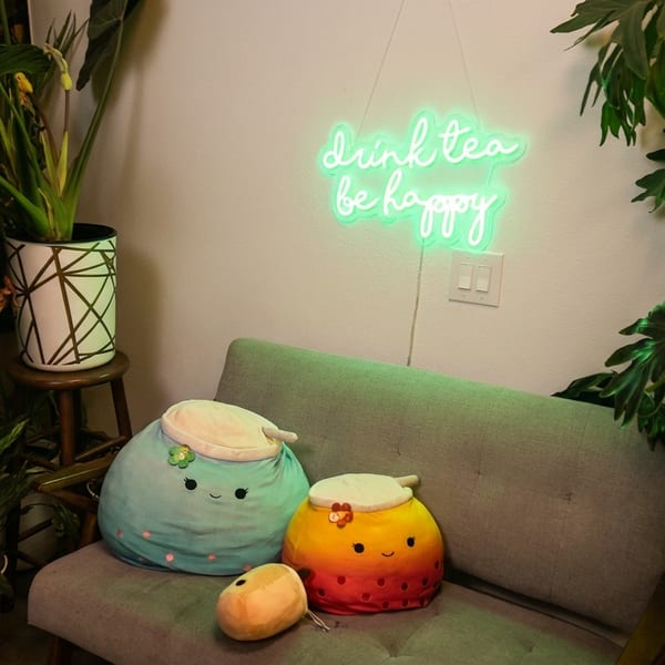 A cozy corner with a gray couch, plush boba tea pillows, plants, and a neon "drink tea be happy" sign.