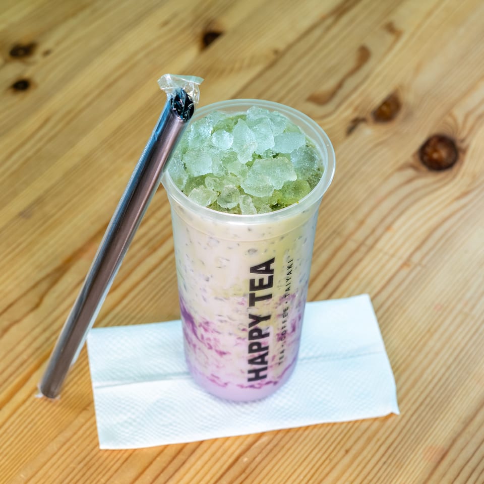 Layered purple and green iced drink in a plastic cup with a metal straw, resting on a napkin atop a wooden table.