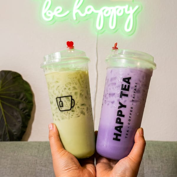 Hands holding two Happy Tea drinks, one green and one purple, under a neon "be happy" sign.
