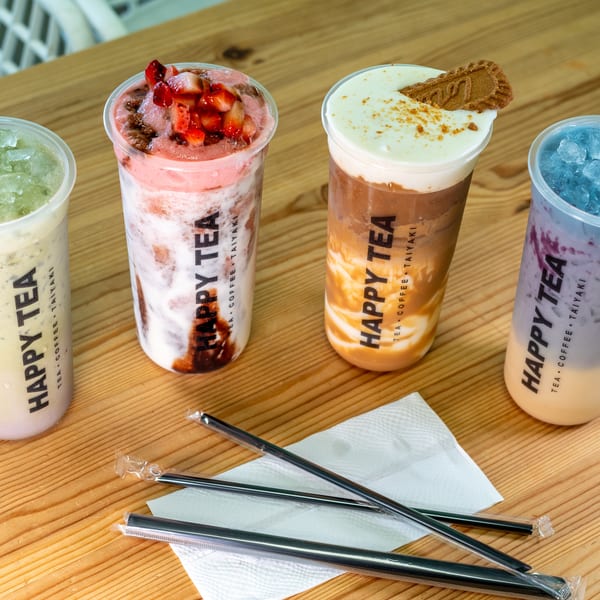 Four colorful Happy Tea drinks, including strawberry, coffee, and blue raspberry, on a wooden table with straws.
