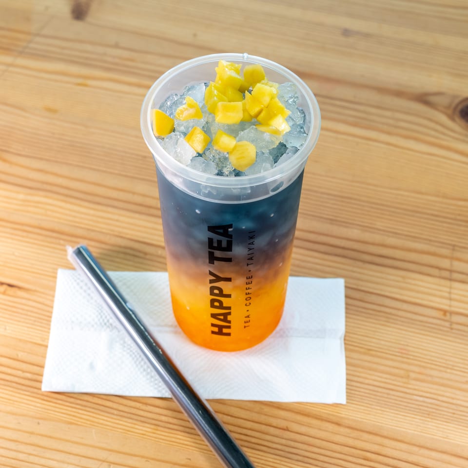 Iced orange tea topped with mango pieces in a branded plastic cup, served with a straw on a napkin-covered wooden table.
