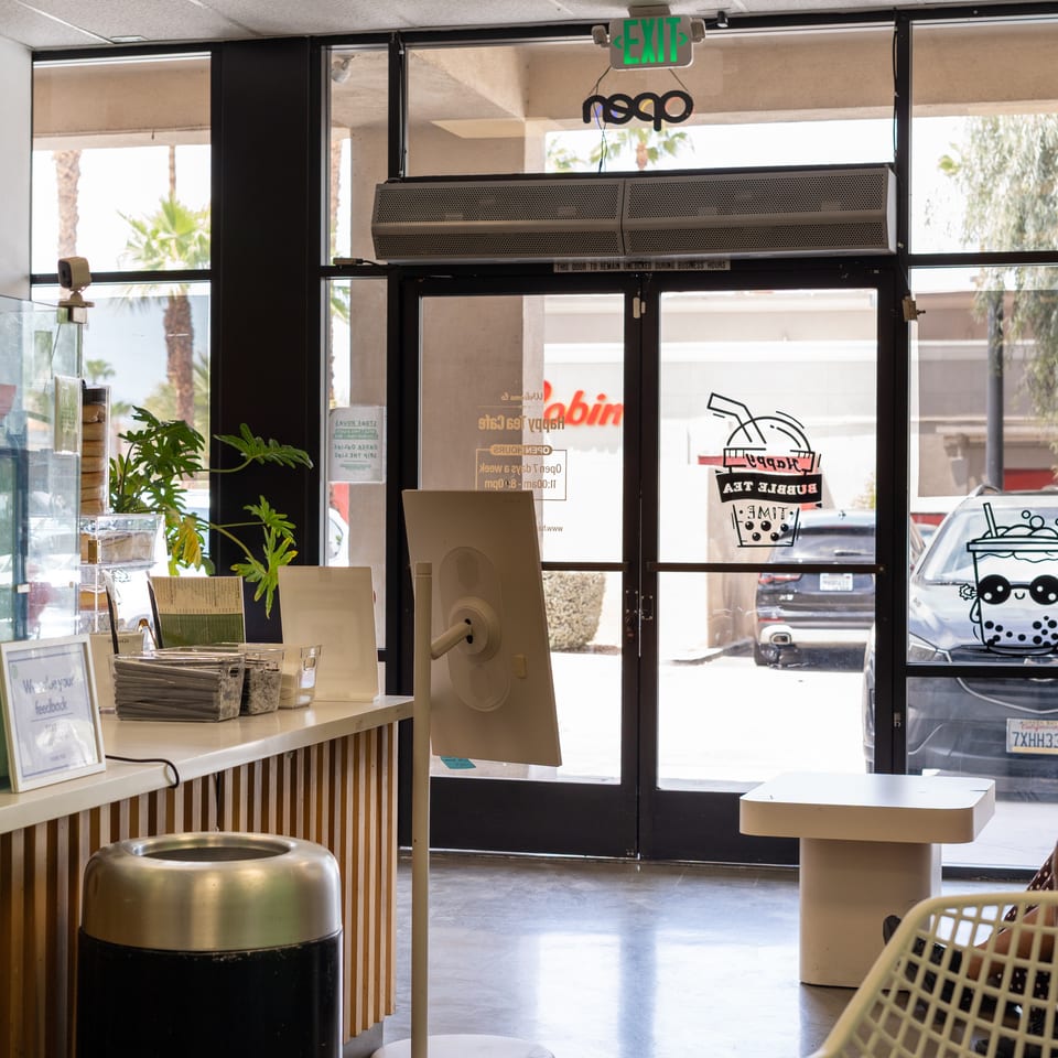 Bright bubble tea shop interior with counter, seating, plants, exit doors, and bubble tea decals on the windows.