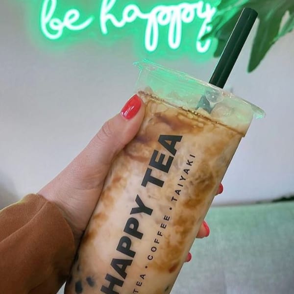 Hand holding a Happy Tea bubble tea with a green neon sign in the background.