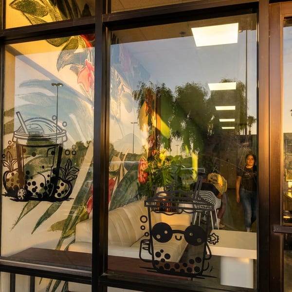 Storefront windows with bubble tea decals, reflecting palm trees and a sunset, with a person inside.