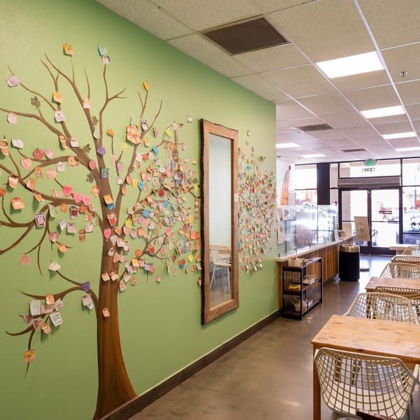 A light green wall with a painted tree covered in colorful customer notes and a mirror, next to empty tables and chairs.