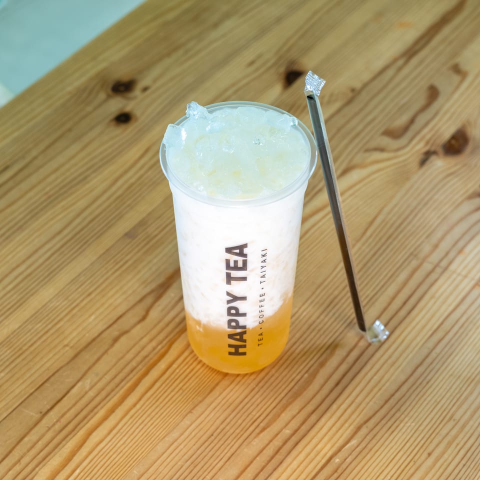 Layered orange iced drink topped with crushed ice in a Happy Tea cup, accompanied by a wrapped metal straw on a wooden table.