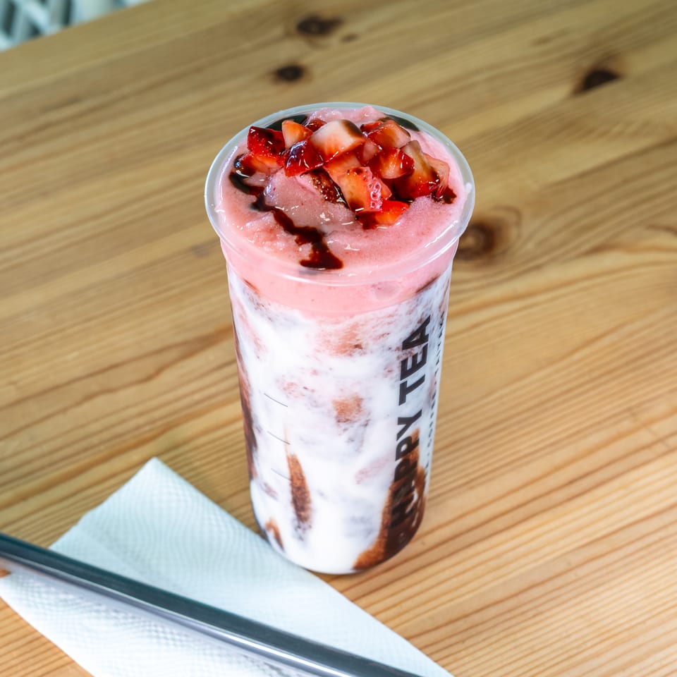 Pink strawberry dessert drink topped with chopped strawberries and chocolate syrup on a wooden table beside a wrapped straw.