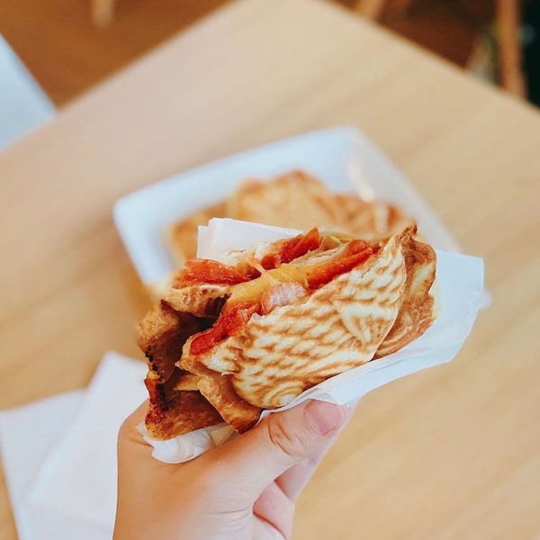 Hand holding a golden-brown, grilled sandwich with red filling, wrapped in white paper.