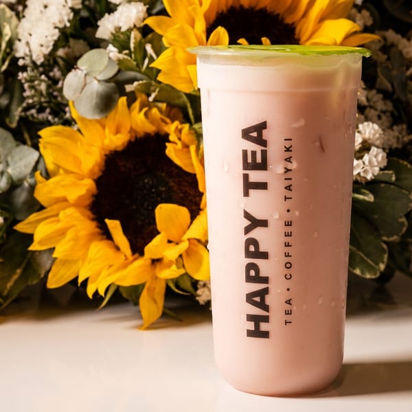 Happy Tea's pink drink with a green lid, next to a bouquet of sunflowers and white flowers.